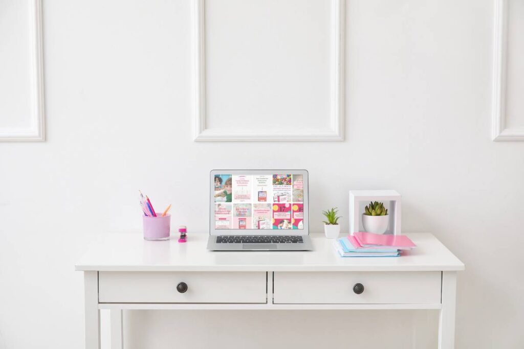 A white desk with a laptop displaying Pinterest website, a cup of pens, a small plant, a pink folder, and stacked notebooks against a white wall.
